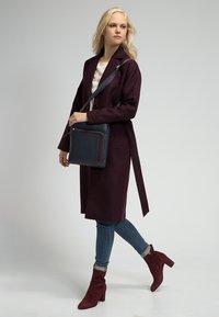 Lungo cappotto bordeaux con cintura, borsa a tracolla in pelle blu navy con zip, jeans e stivali alti bordeaux.