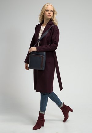 Lungo cappotto bordeaux con cintura, borsa a tracolla in pelle blu navy con zip, jeans e stivali alti bordeaux.