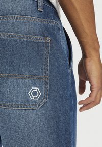 Prim-plan al șoldului unei persoane care poartă blugi din denim albastru, cu un buzunar posterior cusut și un logo geometric alb lângă colțul de jos al buzunarului.