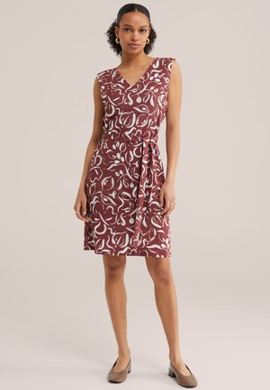 Naine kannab varrukateta burgundia värvi kleiti, millel on valge abstraktne lillemuster ning sobiv vöö, seisab käed kõrval.