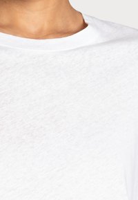 Vit bomullströja med rund hals och strukturerad yta, som visar lätt genomskinlighet och slätt tyg. Närbild av halsringning och axel.