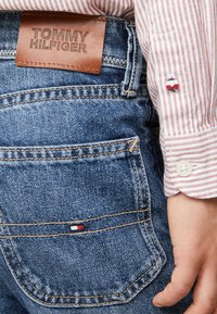 Jeans in denim Tommy Hilfiger con patch in pelle, colore blu medio, taglio dritto, cuciture visibili e un piccolo dettaglio a bandiera sulla tasca.