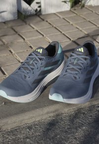 Sportschoenen met een blauw-grijze mesh bovenzijde, donkere accenten en een lichtblauwe zool. Heeft een veterontwerp en een gestructureerd oppervlak voor grip.