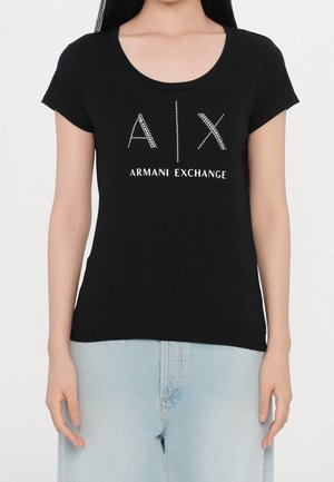 Kobieta ubrana w dopasowaną czarną koszulkę z krótkim rękawem z logo „A|X Armani Exchange” w białe kropki i napis, połączoną z jasnoniebieskimi jeansami.