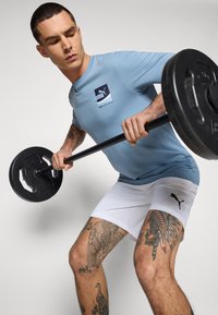 Tricou sport de culoare albastru deschis cu un logo alb și negru, asortat cu șorturi albe ce au un logo negru, ținând o bară cu discuri de greutate.