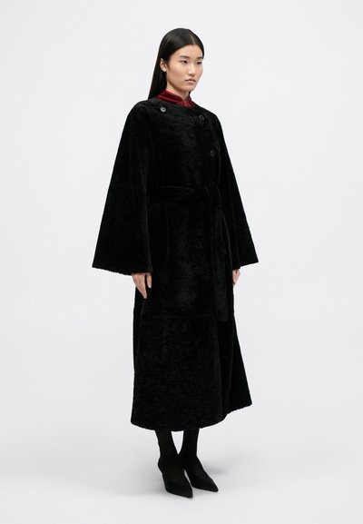 Casaco preto, longo, feito de tecido de pelúcia, com mangas largas, botões duplos e um cinto de tecido. Usado sobre uma camisa vermelha com colarinho e leggings pretas.