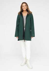 Chaqueta exterior verde, de largo hasta la rodilla, con un alto cuello, cremallera frontal y dos bolsillos laterales. Pantalones blancos y botas de color claro.