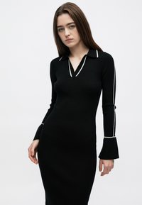 Zwarte geribbelde jurk met een kraag, voorzien van een witte bies langs de halslijn en mouwen. Aansluitende pasvorm met uitlopende manchetten bij de polsen.