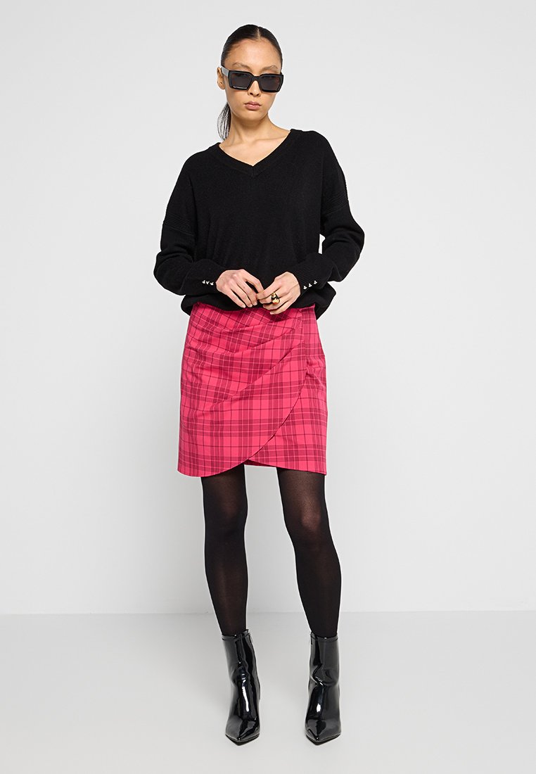 Donna che indossa un maglione nero a V, una gonna avvolgente a quadri rossi, collant neri, stivaletti neri lucidi alla caviglia e occhiali da sole scuri, in piedi contro un muro bianco.