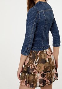 Jeansjacka med en figursydd design, prydd med guldnitar, 3/4-ärmar och en mörkblå tvätt, kombinerad med en blommig volangkjol.