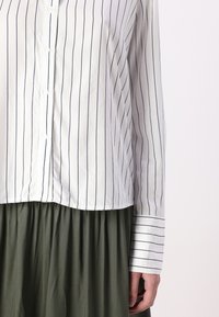 Chemise blanche à boutons avec des rayures verticales navy, dotée d'un ourlet arrondi et de manches longues, portée par-dessus une jupe verte. Texture de tissu lisse.