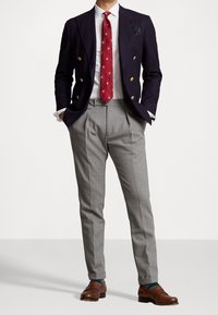 Granatowy dwurzędowy żakiet z złotymi guzikami, szare plisowane spodnie, czerwony krawat w wzory oraz brązowe skórzane loafersy. Pod spodem biała koszula.