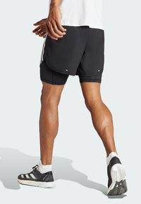 Pantalones cortos deportivos negros con un diseño en capas, que presentan franjas laterales blancas y una textura suave. Combinados con zapatillas negras y blancas, mostrando las piernas.