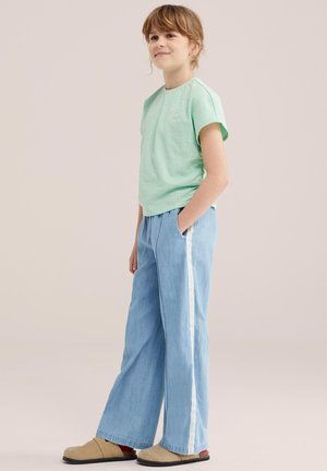 Niño con cabello castaño claro sonriendo, con una camiseta de manga corta color verde menta, pantalones anchos azul claro con rayas blancas en los lados y zapatos beige.