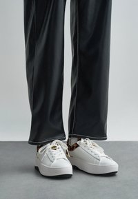 Ténis plataforma brancos com detalhes em padrão de leopardo e ferragens douradas. Material liso; bico arredondado; costura tonal; combinados com calças pretas de couro sintético.