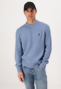 Lichtblauwe geribde trui met een ronde hals, met een klein logo-embleem in rood, wit en blauw. Ggedragen met lichte spijkerbroeken.