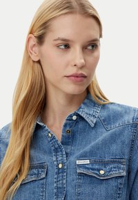 Blonde woman with long hair wearing a blue denim button-up shirt, looking slightly to her right against a plain background.