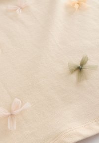 Tissu beige à la texture douce présentant de petits nœuds en tulle rose clair et vert, espacés uniformément sur toute la surface.