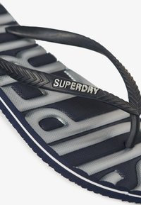 Tongs bleu marine avec une sangle noire texturée arborant le mot 'SUPERDRY' en lettres argentées. La semelle présente un motif géométrique et un accent blanc.