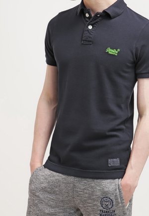 Mand iført sort poloshirt med grønt logo og grå joggingbukser med Franklin & Marshall-logo, hænderne i lommerne, står mod en hvid baggrund.