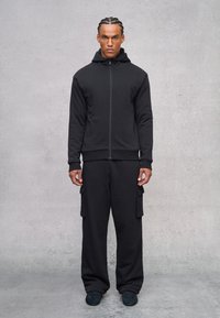 Zwarte zip-up hoodie met een hoge kraag, gecombineerd met losse zwarte cargobroek met zijzakken. Soepele stof met een matte afwerking.