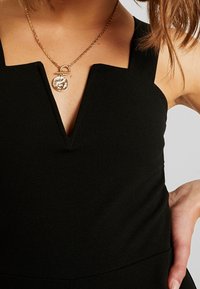 Robe noire avec un décolleté en V et des bretelles larges. Collier en chaîne dorée avec un pendentif circulaire au détail complexe. Tissu doux et texturé.