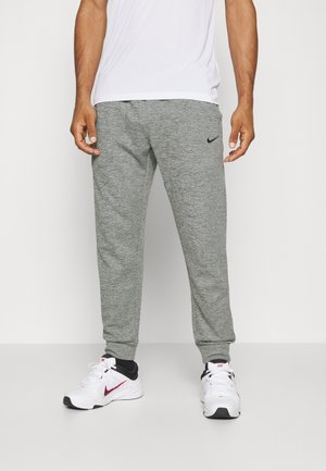 Homme portant un jogging gris Nike, des baskets blanches avec des logos Nike rouges et une chemise blanche sur un fond blanc uni.
