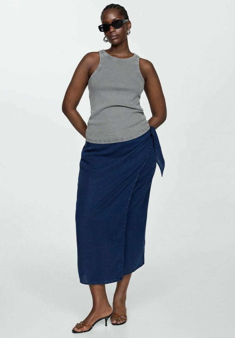 Débardeur gris sans manches associé à une jupe en denim bleu marine avec un nouage sur le côté. Le mannequin porte des lunettes de soleil noires et des sandales à lanières.