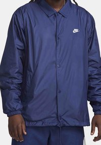 Marinblå vindtät jacka med krage, knappstängning, sidofickor och en liten vit Nike-logotyp på bröstet.