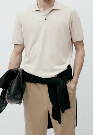 Homme portant un polo beige à manches courtes et un pantalon beige, tenant une veste en cuir noire avec un pull noir noué autour de sa taille.