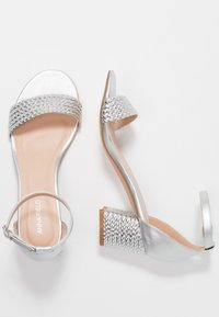 Silver heeled sandals with a braided strap, open toe design, adjustable ankle strap, and a textured block heel. Smooth leather-like finish.