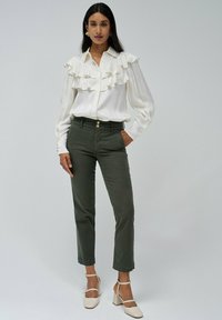 Blusa blanca con detalle de volantes en el frente, combinada con pantalones verdes oliva. La modelo lleva zapatos de tacón bloque claro, que resaltan un diseño casual-chic.