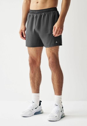 Pantaloni scurți atletici gri, cu un bandă elastică la talie, dintr-un material ușor și cu buzunare laterale. Îmbrăcați cu adidași albi și șosete cu dungi.