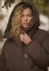 Bruine gewatteerde jas met een grote capuchon, zichtbare knopen en een rits aan de voorkant. Model houdt de kraag vast en draagt een gouden horloge.