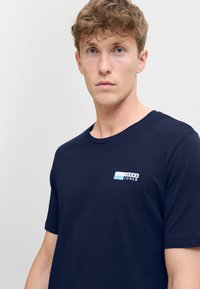 T-shirt di cotone blu navy con maniche corte, con un piccolo logo rettangolare in bianco e azzurro sul lato sinistro del petto.