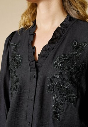 Sort bluse med flæset halsudskæring og knaplukning, der har indviklet sort blomsterbroderi med perlebesætninger foran.