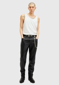 Zwarte leren broek met een kettingdetail, gecombineerd met een witte mouwloze top. Het model staat met de handen in de zakken tegen een neutrale achtergrond.
