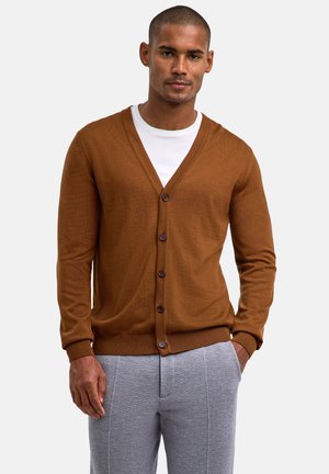 Brauner Strickcardigan mit V-Ausschnitt, Knopfverschluss und gerippten Bündchen. Wird über einem weißen T-Shirt getragen, kombiniert mit grauen Jogginghosen.