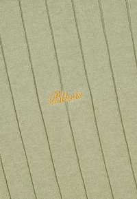 Tissu en maille côtelée vert clair avec une petite broderie cursive orange indiquant « Bikorto » au centre de l'image.