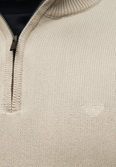 Beež kudumiga kampsun, millel on osaliselt avatud lukuga krae ning rinnal reljeefne logo.