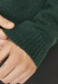 Ciemnozielony sweter z dzianiny z prążkowanym mankietem, prezentujący miękką teksturę i dopasowany krój wokół nadgarstka, z widocznymi detalami dłoni.