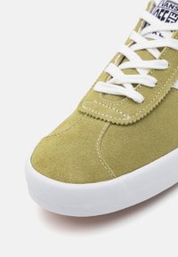 Suede green sneaker with white laces, textured upper, exposed stitching, and a flat white rubber sole. Subtle branding on the tongue.