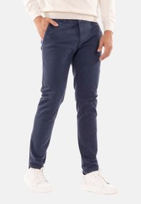 Pantaloni in cotone blu navy con vestibilità slim, dotati di chiusura con zip, tasche laterali e una leggera texture. Abbinati a sneaker bianche.