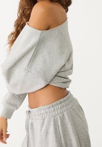 Sweat-shirt gris clair à épaules dénudées en matériau doux, assorti à un pantalon de survêtement à cordon de serrage. Coupe décontractée, poignets et taille côtelés.
