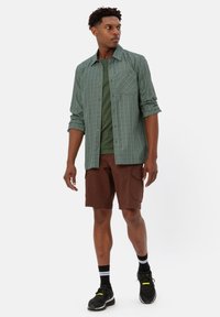 Groene geruite button-up shirt over een donkergroen T-shirt, gecombineerd met bruine cargoshorts; zwarte sportschoenen en gestreepte sokken.