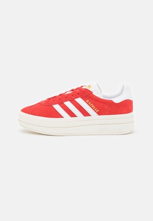 Punainen Adidas Gazelle -lenkkari, jossa valkoiset raidat, valkoinen pohja ja valkoinen kantakiinnike, sivulta kuvattuna yksinkertaisella taustalla.