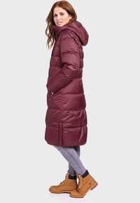 Bordeauxfarbene gepolsterte Jacke mit Kapuze, die ein gestepptes Design, Seitentaschen und einen knielangen Schnitt aufweist. Kombiniert mit grauen Jeans und braunen Stiefeln.