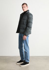 Giacca a vento in grigio scuro, con chiusura a zip e texture trapuntata. Abbinata a jeans azzurri e sneakers nere con suola spessa.