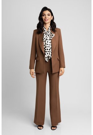 Kobieta o ciemnych włosach, ubrana w brązowy dopasowany garnitur, białą bluzkę z dużymi czarnymi groszkami i czarne buty na obcasie z odkrytymi palcami, stojąca na jasnym tle.