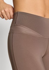 Leggings maro cu talie înaltă, fabricate dintr-un material elastic cu o textură netedă și cusături plate pentru confort. Vedere detaliată a taliei.